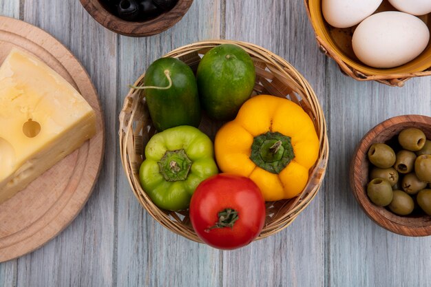 Vista superior de pimientos de colores con pepinos y tomates en canasta con huevos de gallina, queso y aceitunas sobre fondo gris