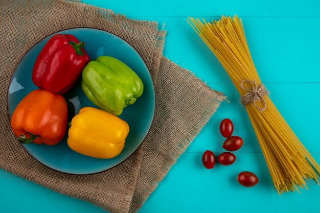 Vista superior de pimientos de colores en una placa azul con pasta cruda y tomates cherry sobre una superficie turquesa