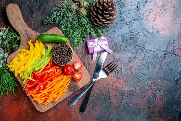 Vista superior de los pimientos cortados de colores, pimienta negra, tomates, pepino, en la tabla de cortar, regalo pequeño, tenedor y cuchillo, ramas de pino en la mesa de color rojo oscuro, espacio libre