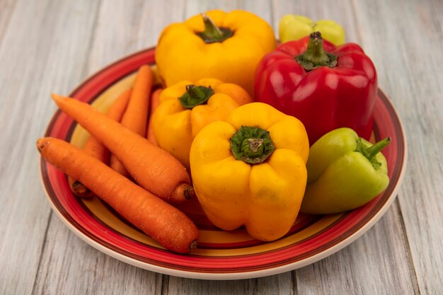 Vista superior de pimientos frescos coloridos en un plato con zanahorias sobre una superficie de madera gris