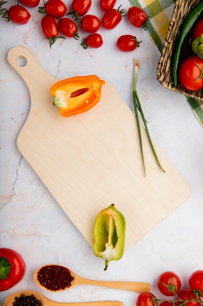 Vista superior de pimientos en rodajas con cebolleta y otras verduras y tabla de cortar sobre superficie blanca