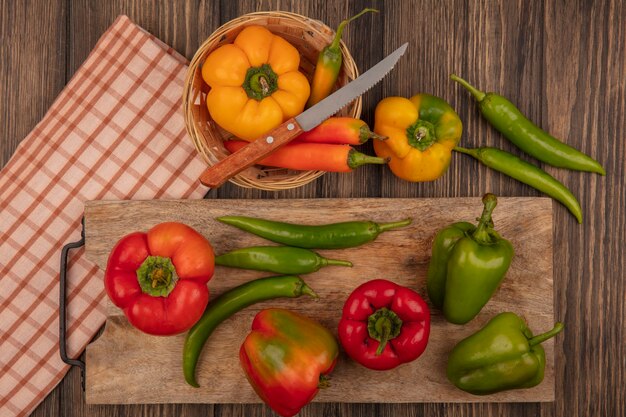 Vista superior de pimientos rojos y verdes frescos en una tabla de cocina de madera con pimientos amarillos en un cubo con un cuchillo sobre un paño marcado en una pared de madera