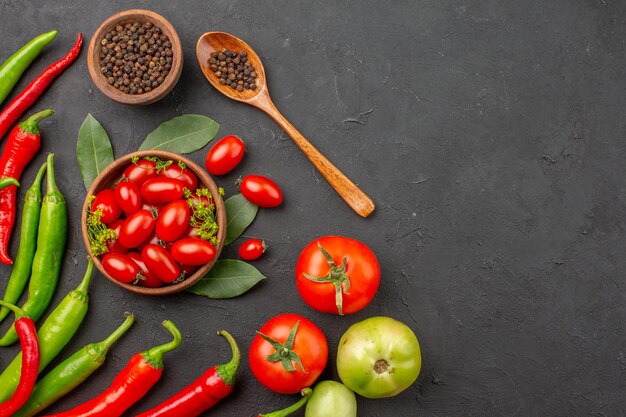 Vista superior de pimientos rojos y verdes y un tazón de tomates cherry, pimienta negra y tomates rojos y verdes en el lado izquierdo de la superficie negra