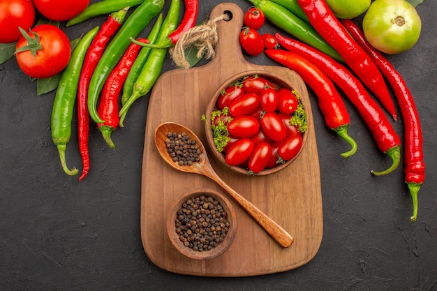 Vista superior, pimientos rojos y verdes, tazones de tomates con tomates cherry y pimienta negra y una cuchara en una tabla de cortar sobre suelo negro con espacio libre