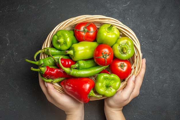 Vista superior de pimientos y tomates en un recipiente de cesta de mimbre en mano femenina sobre superficie oscura