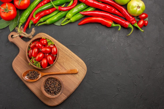 Vista superior de pimientos y tomates rojos y verdes calientes, hojas de laurel, cuencos con tomates cherry y pimienta negra y una cuchara en una tabla de cortar sobre un suelo negro con espacio libre