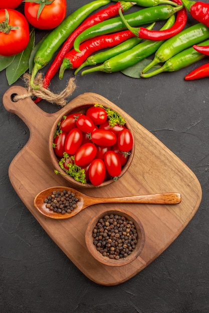 Vista superior de pimientos y tomates rojos y verdes calientes, hojas de laurel, cuencos con tomates cherry y pimienta negra y una cuchara en una tabla de cortar sobre suelo negro