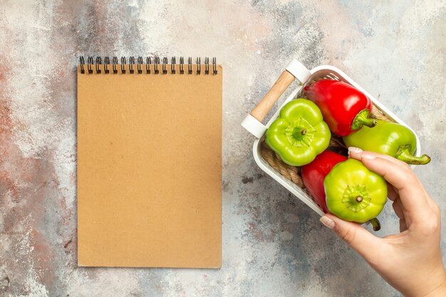 Vista superior de pimientos verdes y rojos en canasta de plástico pimiento verde en mano femenina un cuaderno sobre superficie desnuda