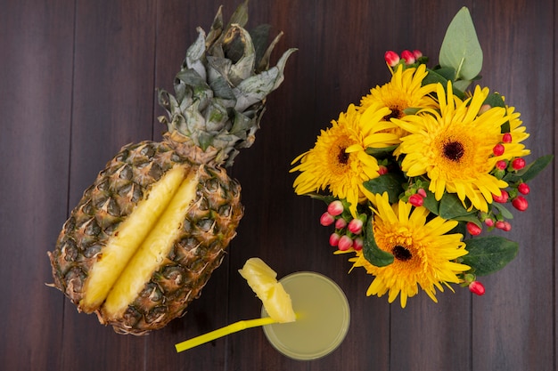 Vista superior de piña y jugo de piña en vidrio con tubo para beber y flores sobre superficie de madera