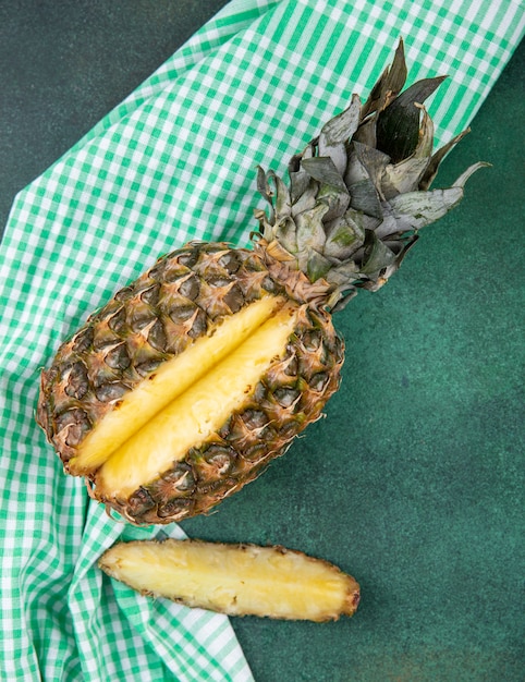 Vista superior de piña con una pieza cortada de fruta entera sobre tela escocesa y superficie verde