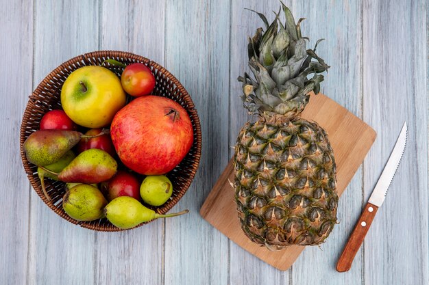 Vista superior de la piña en la tabla de cortar con granada, durazno, manzana, ciruela en la cesta con un cuchillo sobre una superficie de madera