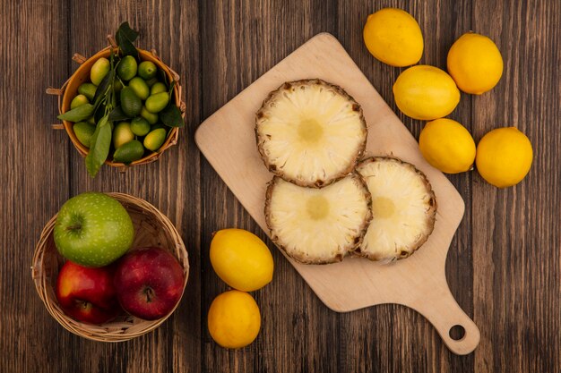 Vista superior de piñas frescas en una tabla de cocina de madera con kinkans en balde con coloridas manzanas en un balde con limones aislado en una pared de madera