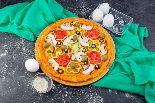 Vista superior de la pizza de champiñones con tomates, aceitunas, champiñones, todos en rodajas por dentro con harina sobre el fondo gris, masa de pizza de tejido verde italia