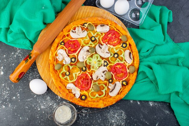 Vista superior de la pizza de hongos con tomates, aceitunas, champiñones, todos en rodajas por dentro con harina en el escritorio gris, masa de pizza de tejido verde, comida italiana