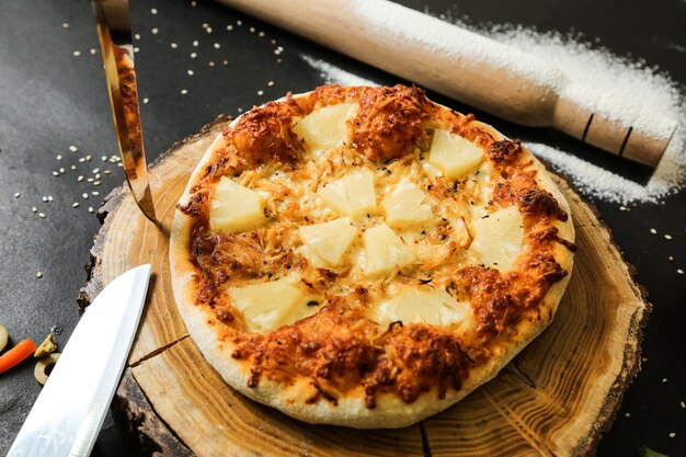 Vista superior pizza de piña en el stand con cuchillos rodillo y harina en la mesa negra