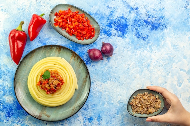 Vista superior de una placa gris con deliciosos espaguetis picados y cebollas de pimiento rojo entero y mano sosteniendo la carne en la mesa azul