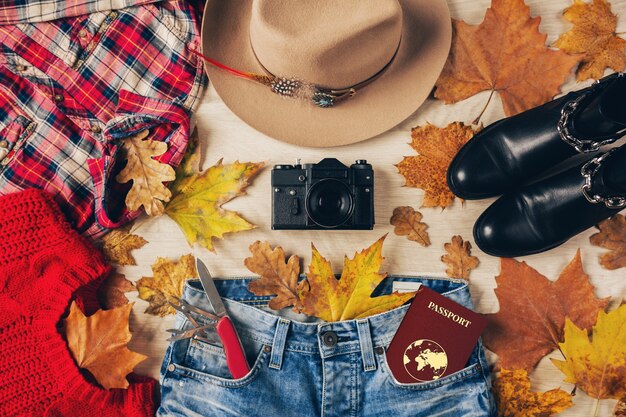 Vista superior en plano de accesorios de estilo de mujer, suéter rojo, camisa de franela a cuadros, jeans, botas de cuero negro, tendencia de moda otoñal, cámara de fotos vintage, cuchillo suizo, pasaporte, traje de viajero