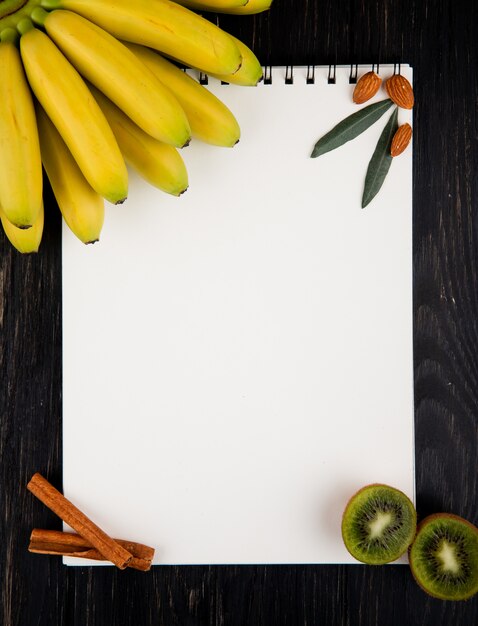 Vista superior de plátanos frescos y kiwis con un cuaderno de dibujo, palitos de almendras y canela en negro