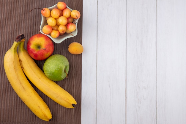 Foto gratuita vista superior de plátanos con manzanas y cerezas blancas en una servilleta marrón sobre una superficie blanca