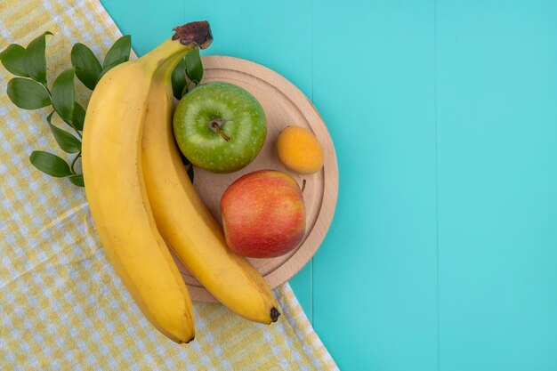 Vista superior de plátanos con manzanas en un soporte sobre una toalla a cuadros amarilla sobre una superficie azul