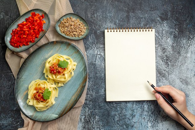 Vista superior de un plato azul con una deliciosa comida de pasta servida con tomate y carne para la cena en una toalla de color canela con sus ingredientes mano sosteniendo un bolígrafo en un cuaderno de espiral