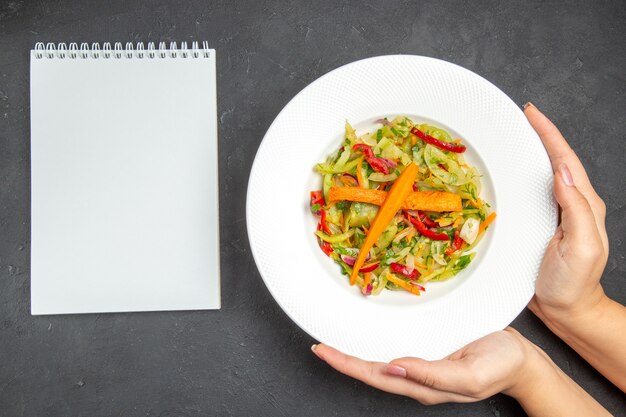 Vista superior del plato de ensalada de una apetitosa ensalada con verduras en las manos cuaderno blanco