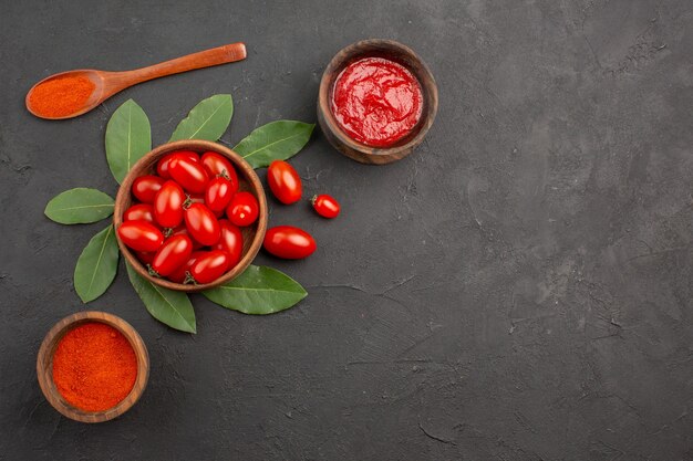 Vista superior de un plato de laurel de tomates cherry deja una cuchara de madera y tazones de salsa de tomate y pimiento picante en polvo sobre la mesa negra