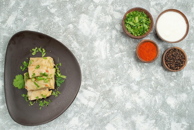 Vista superior desde un plato lejano con hierbas Plato de repollo relleno junto a tazones con hierbas y especias de crema agria de pimienta negra en el lado izquierdo y derecho de la mesa