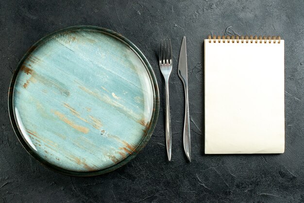 Vista superior del plato redondo de acero tenedor y cuchillo de cena cuaderno sobre mesa negra