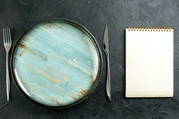 Vista superior plato redondo cena cuchillo y tenedor cuaderno sobre mesa negra