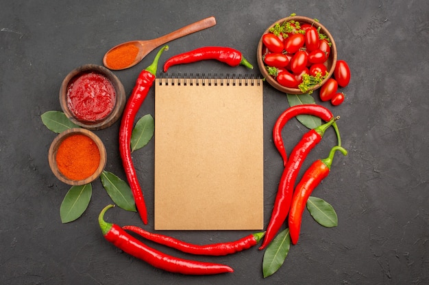 Vista superior de un plato de tomates cherry, pimientos rojos picantes, un cuaderno, una cuchara de madera, hojas de laurel y tazones de salsa de tomate y pimiento picante en polvo sobre la mesa negra