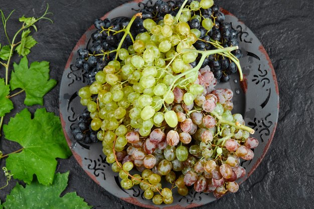 Vista superior de un plato de uvas mixtas alrededor de hojas de parra sobre fondo oscuro. Foto de alta calidad