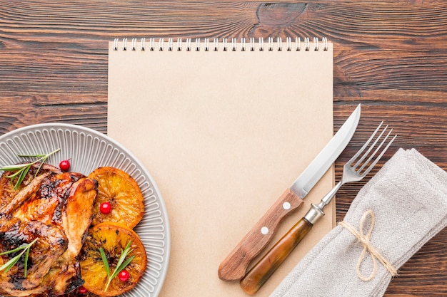 Vista superior de pollo al horno y rodajas de naranja en un plato con cubiertos y cuaderno en blanco