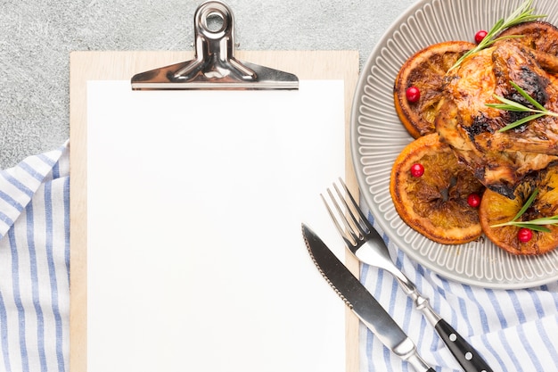 Vista superior de pollo al horno y rodajas de naranja en un plato con papel de cocina y portapapeles en blanco
