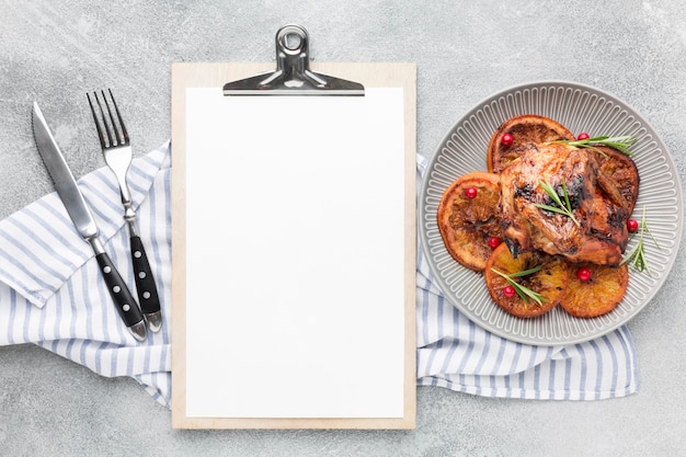 Vista superior de pollo al horno y rodajas de naranja en un plato con una toalla de cocina y un bloc de notas en blanco