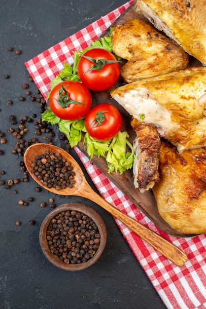 Vista superior de pollo al horno sobre tablero de madera, pimienta negra en un tazón pequeño, cuchara de madera sobre mesa negra