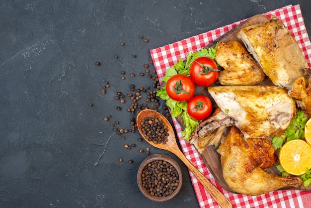 Vista superior de pollo al horno sobre tablero de madera, pimienta negra en un tazón pequeño, cuchara de madera sobre negro