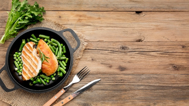 Vista superior de pollo a la parrilla y guisantes en una sartén con cubiertos y espacio de copia