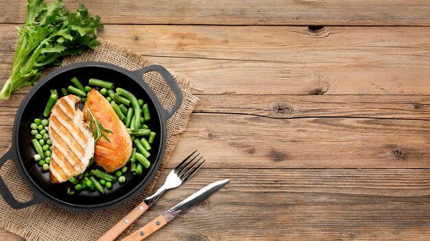 Vista superior de pollo a la parrilla y guisantes en una sartén con cubiertos y espacio de copia