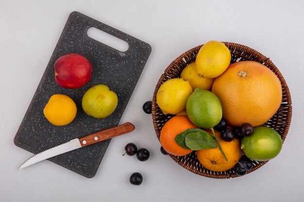 Vista superior de pomelo con naranjas limas y limones en una canasta con ciruelas cereza y melocotones en una tabla de cortar con un cuchillo sobre un fondo blanco.