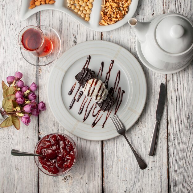 Vista superior postre en placa con té, nueces tetera, mermelada de frutas, flores sobre fondo blanco de madera.