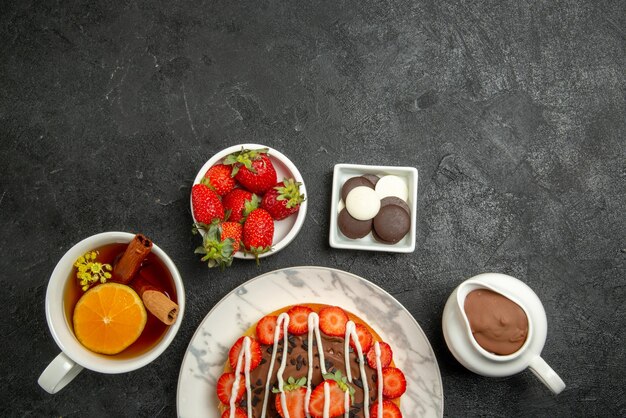 Vista superior de primer plano apetitoso pastel de fresas con crema de chocolate y una taza de té con limón y canela junto a los tazones de fresas con chocolate y crema de chocolate en la mesa oscura