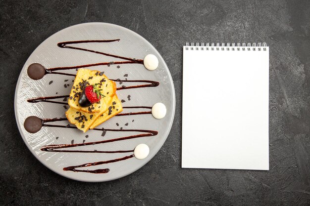 Vista superior de primer plano cuaderno blanco de postre junto a la placa gris de apetitoso pastel con fresas y salsa de chocolate en el lado izquierdo de la mesa oscura