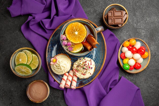 Vista superior de primer plano dulces en la mesa una taza de té con limón canela palitos galletas en el plato cuencos de caramelos de colores rodajas de chocolate de frutas cítricas y crema de chocolate