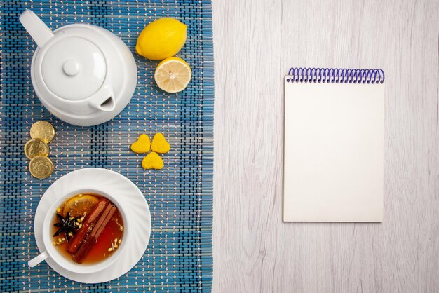 Vista superior de primer plano dulces y una taza de té una taza de té de hierbas con limón y canela limón diferentes dulces tetera sobre el mantel a cuadros junto al cuaderno blanco