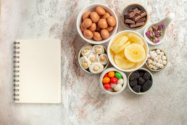 Vista superior de primer plano dulces en tazones cuaderno blanco junto a los tazones de diferentes dulces frutos secos y bayas en la mesa