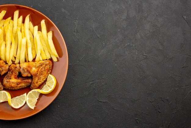 Vista superior de primer plano fastfood naranja plato de papas fritas, alitas de pollo y limón en el lado izquierdo de la mesa oscura