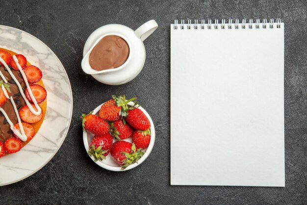 Vista superior de primer plano de fresas con crema de chocolate apetitoso pastel con bayas tazones de cuaderno blanco de fresas y crema de chocolate sobre la mesa
