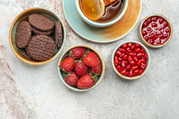 Vista superior de primer plano fresas una taza de té negro con palitos de limón y canela tazones de diferentes bayas galletas de chocolate semillas de granada sobre la mesa