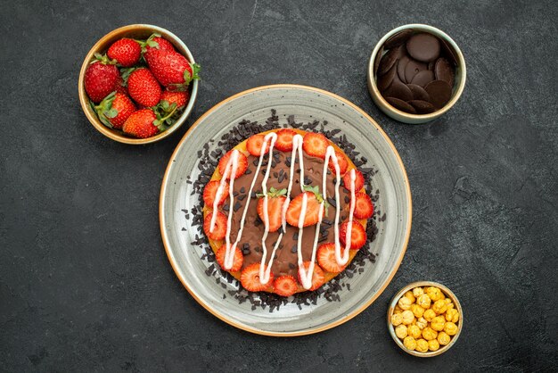 Vista superior de primer plano pastel con chocolate apetitoso pastel con chocolate y fresa y tazones de fresa, avellana y chocolate en el centro de la mesa negra